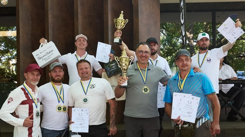 Група чоловіків позує для фото після завершення турніру, тримаючи в руках кубки, медалі на стрічках у кольорах прапора України та почесні дипломи. Нагороджені учасники посміхаються, деякі з них тримають брендовані паперові пакети, а на задньому плані видно колони будівлі та зелені дерева.