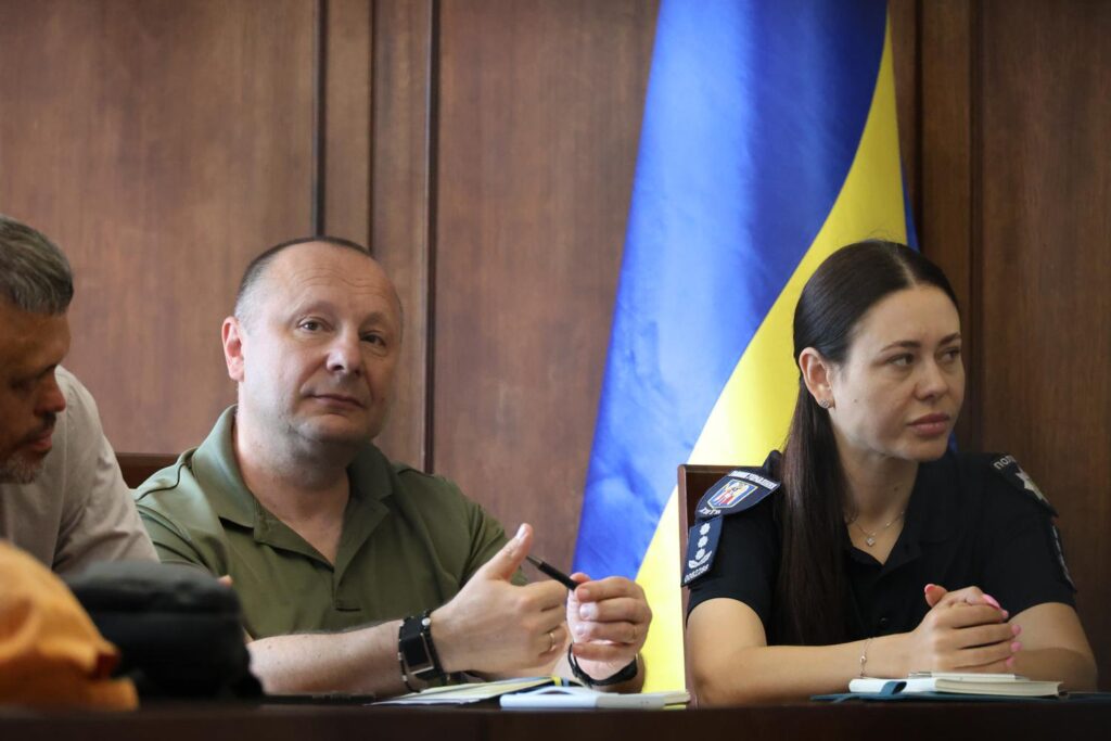 На фото зображено трьох людей за столом під час офіційної зустрічі на тлі великого синьо-жовтого прапора України. У центрі кадру сидить чоловік середнього віку у зеленій сорочці-поло з короткими рукавами. Він тримає в руках ручку, дивиться в об'єктив камери та жестикулює правою рукою під час розмови. Праворуч від нього сидить жінка з темним волоссям, зібраним ззаду, одягнена у чорну форму Національної поліції України з шевронами на рукаві. Вона зосереджено дивиться ліворуч, склавши руки перед собою на столі поруч із блокнотом. З лівого краю фото частково видно третього чоловіка, який нахилився вперед і дивиться вниз. На столі перед присутніми лежать відкриті блокноти.