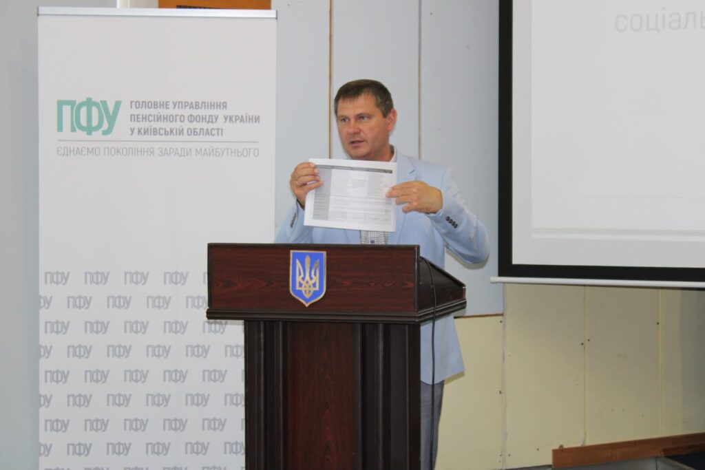Директор Департаменту соціального захисту населення Київської обласної державної адміністрації Ігор Мещан виступає за трибуною з гербом України під час засідання Головного управління Пенсійного фонду України у Київській області. Він демонструє присутнім документ, поруч розташовані брендований банер Пенсійного фонду та екран для презентації.