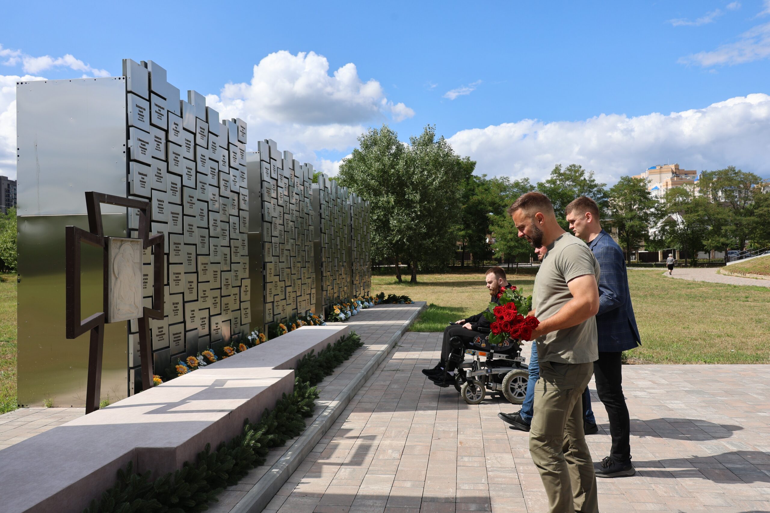 Заступник голови Київської обласної державної адміністрації Іван Добровольський та секретар Бучанської міської ради Тарас Шаправський стоять біля меморіалу на території храму Св. Апостола Андрія Первозванного. Вони тримають букет червоних троянд. На задньому плані видно ветерана Валентина Олексієнка у кріслі колісному в кріслі колісному, а навколо розташована зелена зона з деревами під хмарним синім небом.