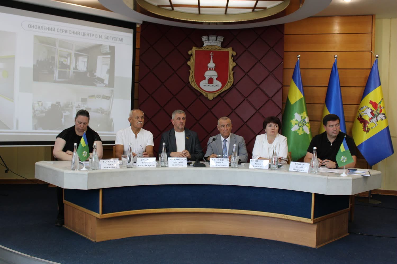 За столом сидять шість учасників засідання колегії Головного управління Пенсійного фонду України у Київській області. На задньому плані розміщені герб міста та три прапори, серед яких Державний прапор України. Ліворуч на екрані демонструється презентація про оновлений сервісний центр у Богуславі. На столі — мікрофони, таблички з іменами учасників, пляшки з водою та настільні прапорці Київської області.