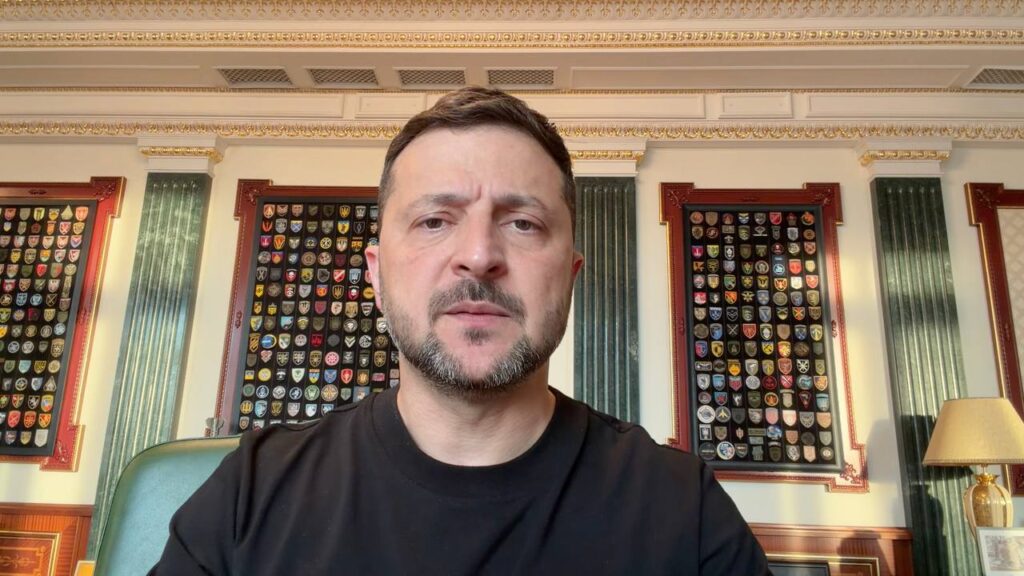 Президент України Володимир Зеленський записує відеозвернення, сидячи за столом у світлому кабінеті. На тлі — великі стенди з військовими шевронами різних підрозділів, зелені мармурові колони та настільна лампа.