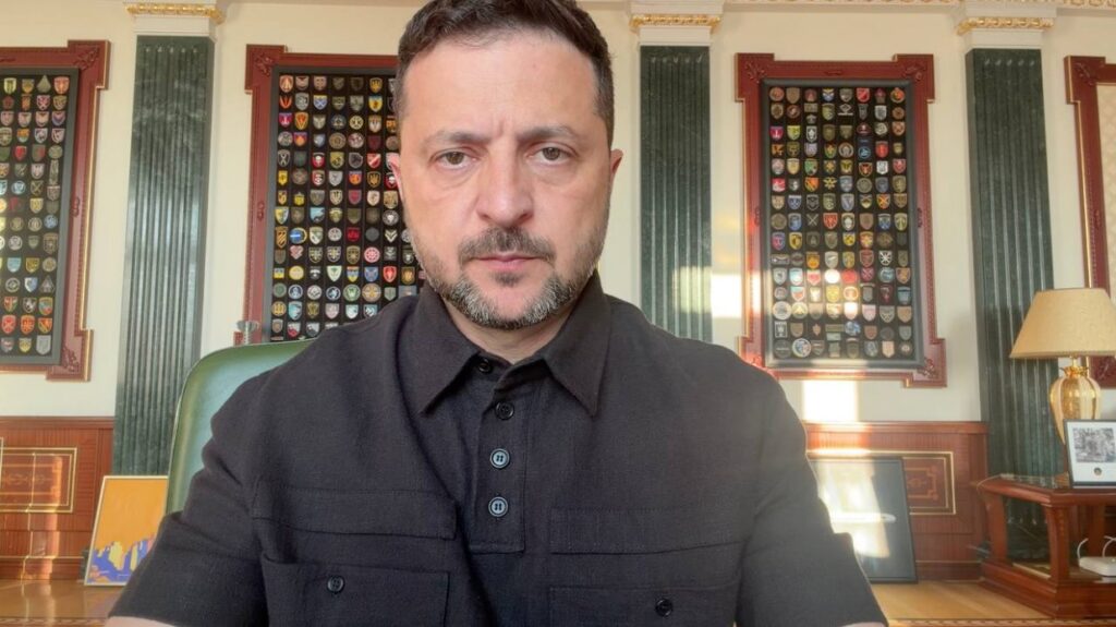 Президент України Володимир Зеленський записує відеозвернення, сидячи за столом у світлому кабінеті. На тлі — великі стенди з військовими шевронами різних підрозділів, зелені мармурові колони та настільна лампа.