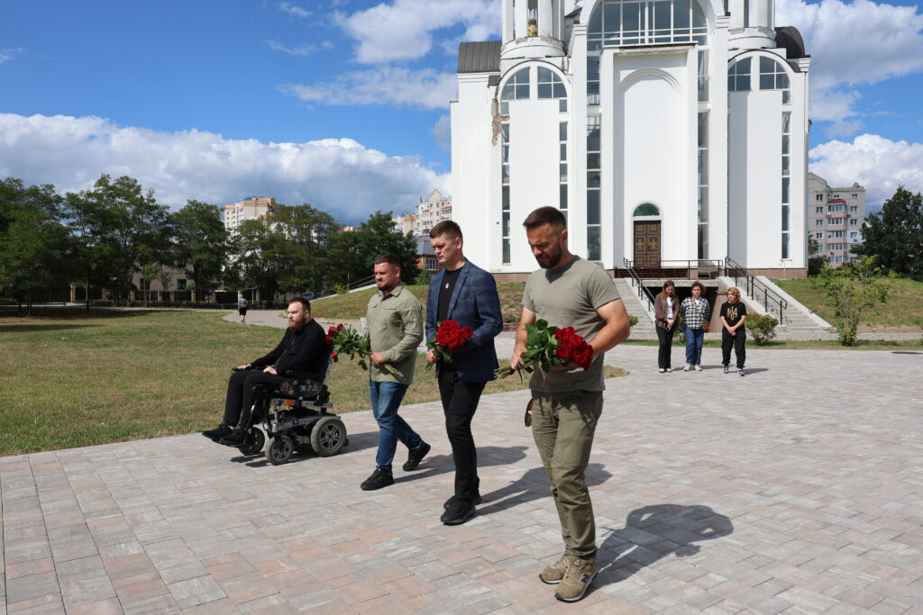 Група людей несе букети червоних троянд, чоловік у кріслі колісному їде брукованою площею на тлі великого білого сучасного храму. Позаду них біля сходів церкви стоять три жінки, а навколо розкинулася зелена зона з деревами під синім небом із хмарами.