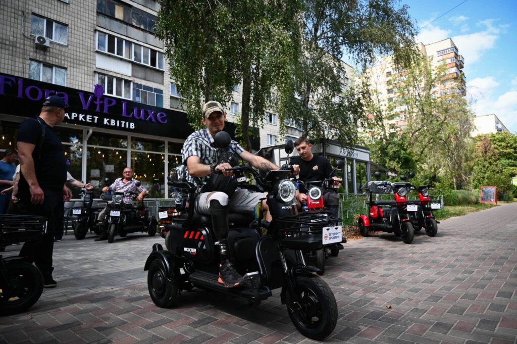 Чоловік з протезом ноги сидить на чорному триколісному електроскутері на міській вулиці з бруківкою, на тлі інших людей та скутерів.