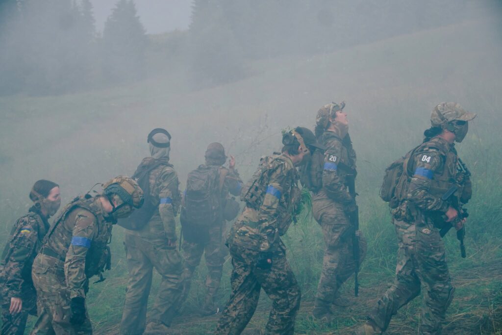 Група людей у військовій формі та захисному спорядженні рухається по трав'янистому схилу крізь густий білий дим або туман. На тлі видно силуети ялин. Деякі учасники мають сині пов'язки на рукавах та тактичні рюкзаки, а один із них тримає зброю.