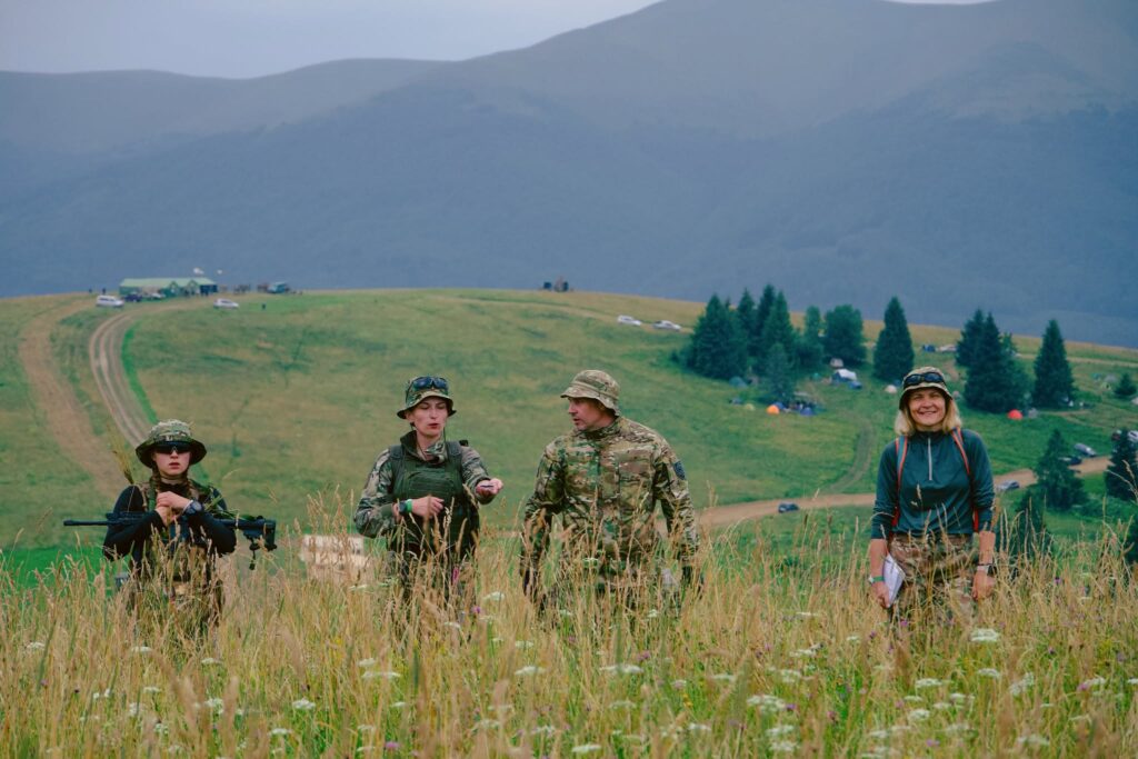 Чотири людини стоять у високій траві на зеленому пагорбі на тлі гірського пейзажу. Троє з них одягнені у камуфляжний одяг та панами, один тримає зброю, а жінка праворуч — у цивільному спортивному одязі та капелюсі. На задньому плані видно намети, автомобілі та поодинокі ялини.