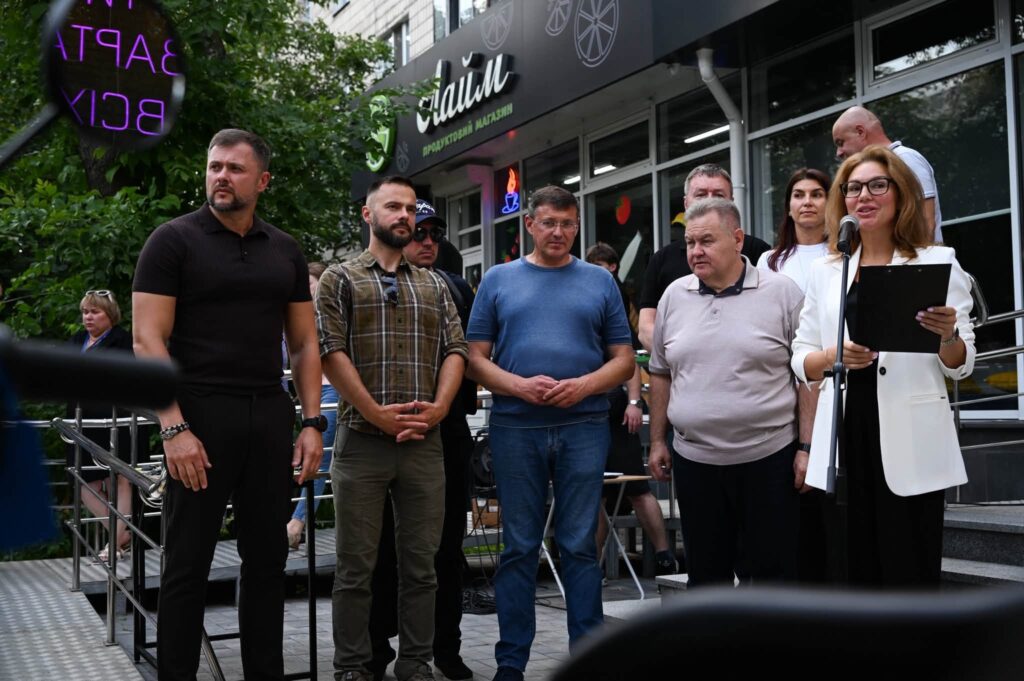 Жінка в окулярах та білому піджаку виступає перед мікрофоном із папкою в руках на тлі групи людей.