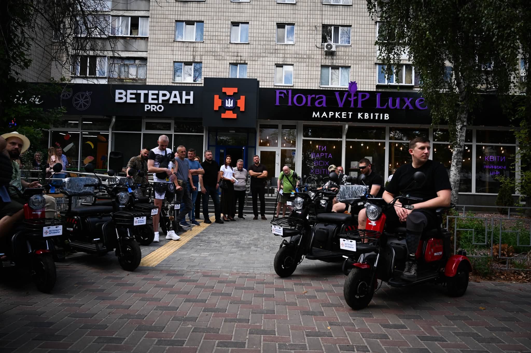 Група людей зібралася на вулиці перед будівлею з вивіскою «Ветеран Про». Декілька чоловіків сидять на триколісних електроскутерах.