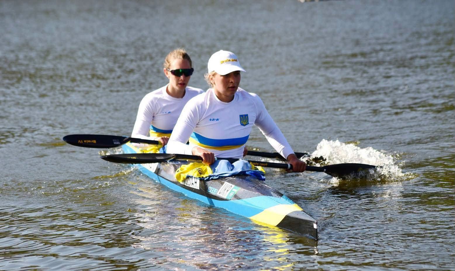 Дві українські веслувальниці у спортивній формі збірної України змагаються на двомісному байдарковому човні. Спортсменки синхронно веслують по воді, а на носі човна видно синьо-жовтий прапор.