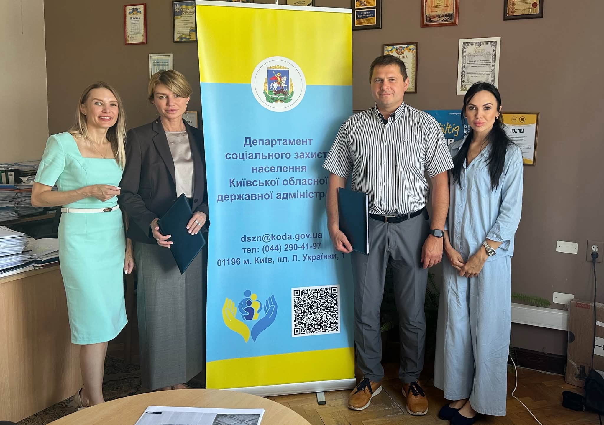 Чотири людини (три жінки та чоловік) стоять у кабінеті поруч із вертикальним жовто-блакитним інформаційним банером Департаменту соціального захисту населення Київської ОДА. Директор Департаменту соціального захисту населення Київської ОДА Ігор Мещан та виконавча директорка міжнародного благодійного фонду «Українська фундація громадського здоров’я» Галина Скіпальська тримають у руках папки з документами.