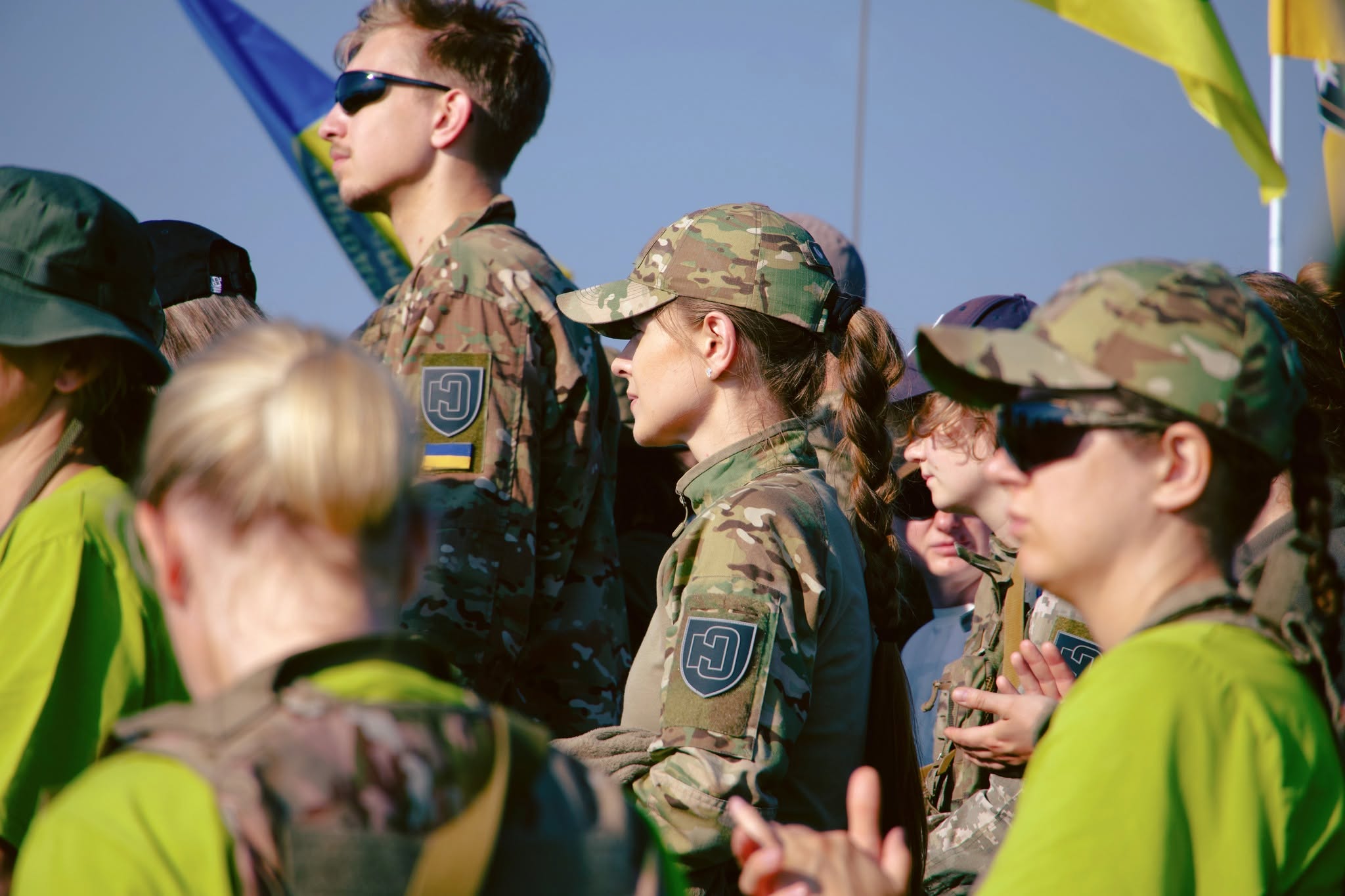 Група молодих людей стоїть у профіль та анфас просто неба під яскравим сонцем. У центрі крупним планом зображена дівчина з довгим волоссям у камуфляжній кепці та формі, на плечі якої прикріплений шеврон Київського обласного центру підготовки населення до національного спротиву з літерами «Н» та «С». Поруч із нею стоять інші учасники. Деякі з них у сонцезахисних окулярах та яскраво-зелених футболках. На розмитому задньому плані видно синьо-жовті прапори на тлі світлого неба.