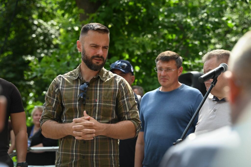 Заступник голови Київської обласної державної адміністрації Іван Добровольський стоїть із зчепленими руками перед мікрофоном на тлі інших людей та зелених дерев.