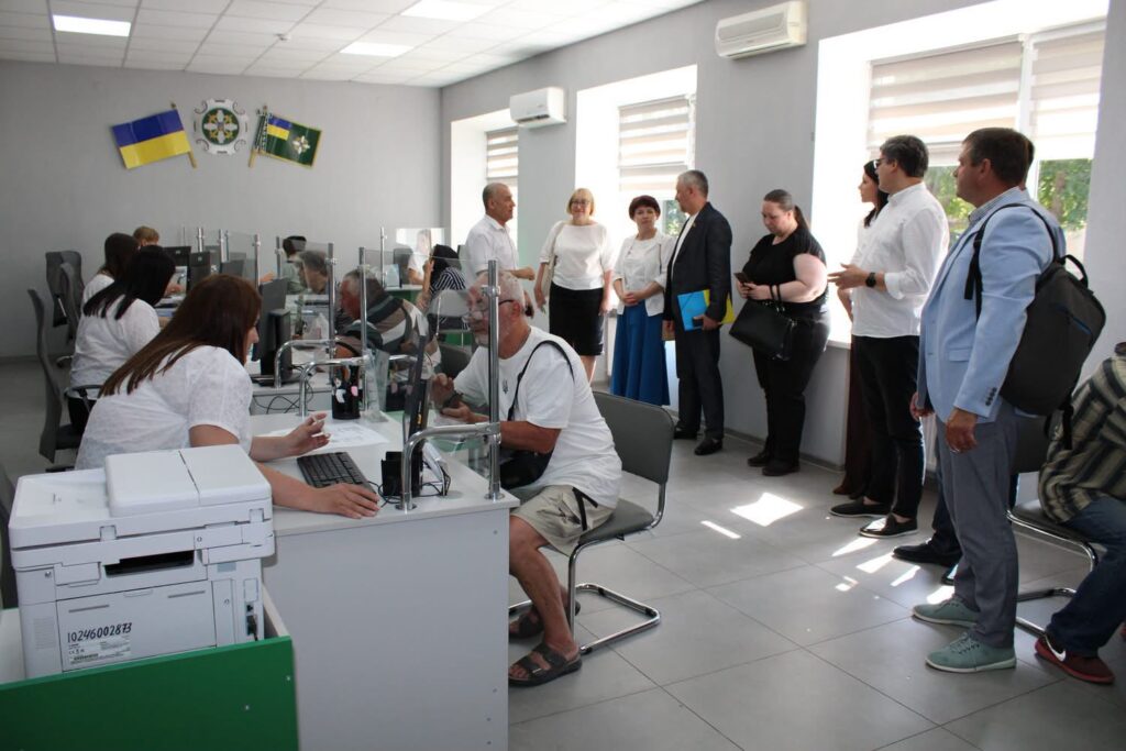 У сучасному сервісному центрі працівники обслуговують відвідувачів за робочими місцями, відокремленими прозорими захисними екранами. Праворуч група людей оглядає роботу установи. На стіні розміщені державний прапор України, емблема установи та прапор громади, приміщення добре освітлене денним світлом.