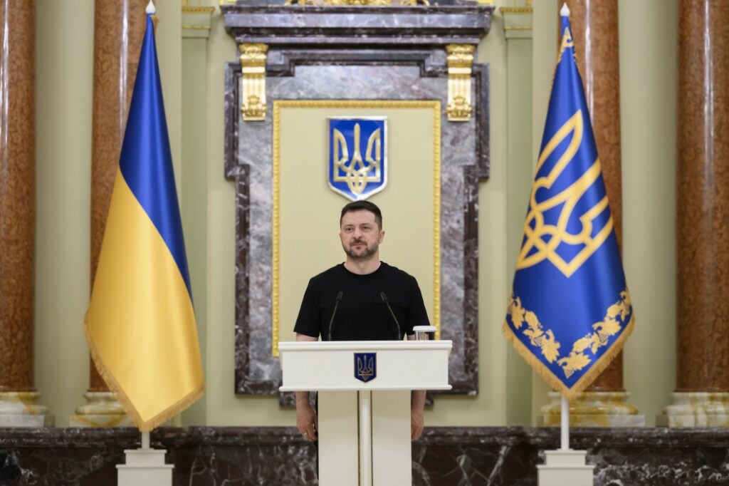 Президент України Володимир Зеленський стоїть за білою трибуною з малим гербом України по центру під час офіційного виступу. Він одягнений у чорну футболку, а позаду нього на стіні розміщено великий синьо-жовтий державний герб України в золотій рамці. Обабіч трибуни встановлено два великі прапори: ліворуч — державний прапор України, праворуч — синій штандарт Президента з золотим тризубом та орнаментом.