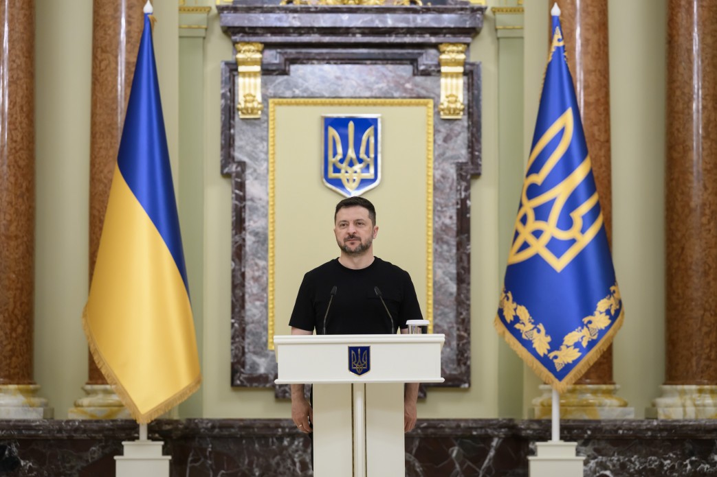 Президент України Володимир Зеленський стоїть за білою трибуною з малим гербом України по центру під час офіційного виступу. Він одягнений у чорну футболку, а позаду нього на стіні розміщено великий синьо-жовтий державний герб України в золотій рамці. Обабіч трибуни встановлено два великі прапори: ліворуч — державний прапор України, праворуч — синій штандарт Президента з золотим тризубом та орнаментом.