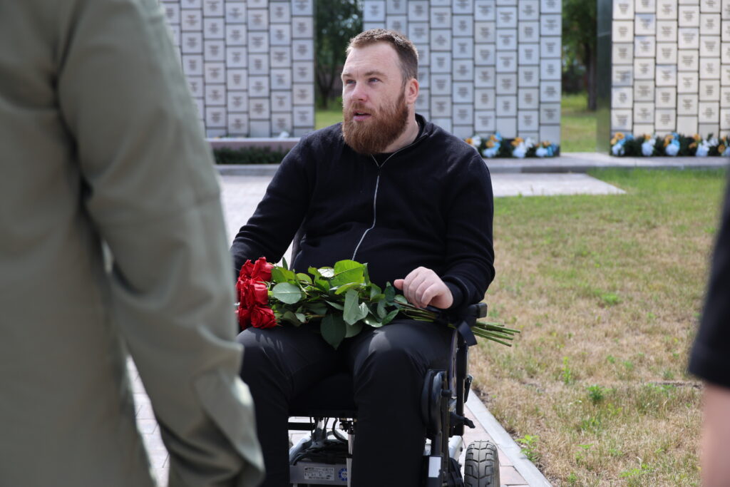 Керівник Бучанського ветеранського простору «Ветеран ПРО», учасник бойових дій Валентин Олексієнко у кріслі колісному тримає букет червоних троянд. Він знаходиться у Бучі на території храму Святого Апостола Андрія Первозванного. На задньому плані видно стели з написами.