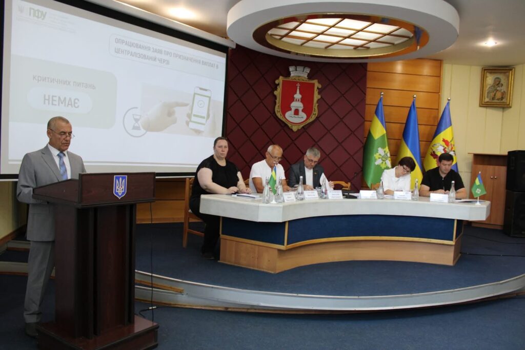Засідання колегії Головного управління Пенсійного фонду України: доповідач виступає за трибуною, поруч за столом президії сидять учасники наради. На екрані демонструється презентація про централізоване опрацювання заяв, позаду розміщені прапори України та громади, а також герб Переяслава.