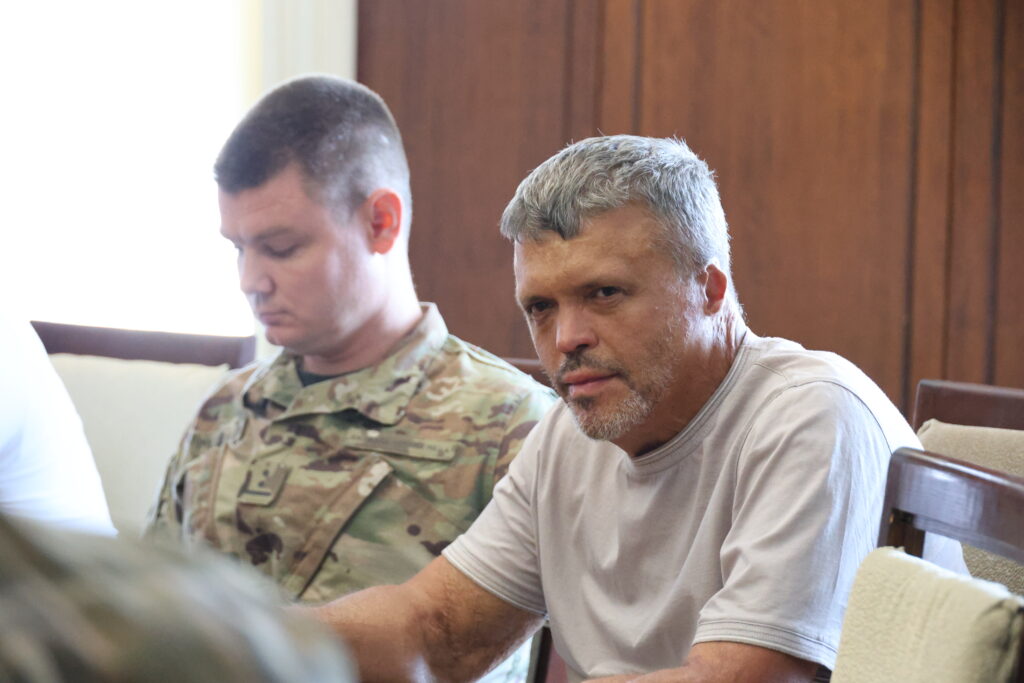Двоє чоловіків сидять поруч у приміщенні під час офіційного заходу. На передньому плані праворуч сидить чоловік середнього віку із сивіючим коротким волоссям, легкою сивою борідкою та вусами. Він одягнений у просту світло-сіру футболку та дивиться прямо в об'єктив камери із серйозним, злегка примруженим поглядом. Ліворуч від нього, трохи на задньому плані, сидить молодий військовослужбовець у камуфляжній формі із шевроном на грудях. Його голова опущена, а погляд спрямований донизу, ніби він читає або робить записи. Тло за ними складається з гладкої дерев'яної стіни коричневого кольору, а зліва присутнє м'яке світло від вікна.
