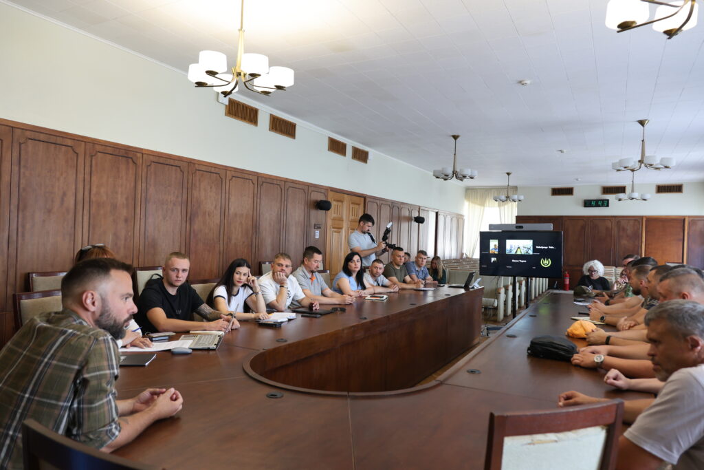 У просторій залі засідань за великим U-подібним дерев'яним столом сидять понад двадцять людей. Учасники зустрічі — переважно чоловіки та жінки молодого й середнього віку, одягнені у повсякденний та напівформальний одяг (футболки, сорочки). Деякі з них роблять записи або дивляться на протилежний бік столу. На дальньому кінці столу встановлено великий телевізійний екран, на якому відображається інтерфейс онлайн-конференції (відеодзвінка) з кількома вікнами учасників. Стіни кімнати оздоблені класичними дерев'яними панелями коричневого кольору, а на білій стелі розміщені великі підвісні люстри з кількома плафонами. На столі перед людьми лежать блокноти, ручки, телефони та ноутбуки.