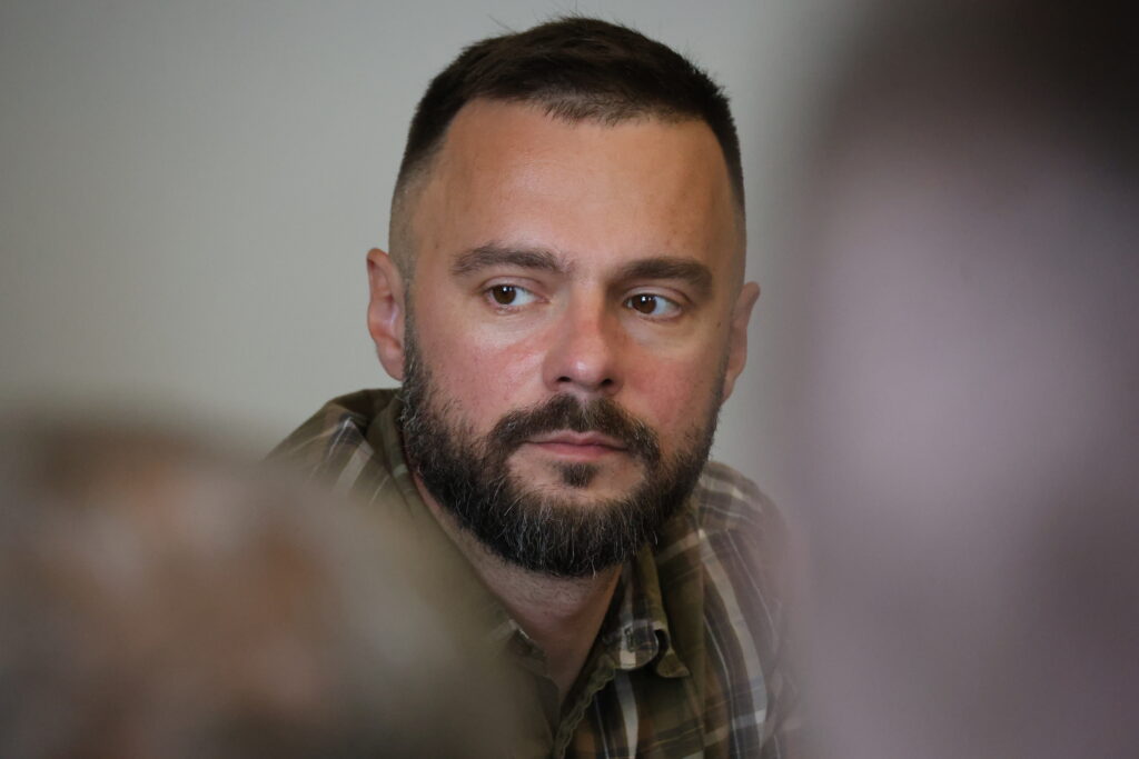 На фото — заступник голови Київської обласної державної адміністрації, ветеран Іван Добровольський. Він одягнений у сорочку у зелено-коричневу клітинку та дивиться кудись убік, ліворуч від камери. Його вираз обличчя зосереджений, серйозний та уважний. Тло за ним світле і однотонне, але передній план зліва та справа частково перекритий сильно розмитими силуетами голів інших людей, що створює ефект глибини кадру та фокусує увагу суто на обличчі чоловіка.