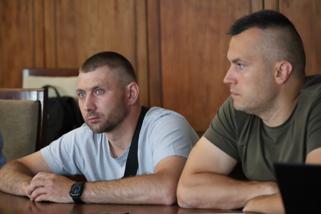 Двоє чоловіків із короткими стрижками сидять поруч за столом під час «круглого столу».Чоловік ліворуч одягнений у світло-сіру футболку, має легку щетину та виразні блакитні очі. Через його плече перекинуто чорний ремінь сумки, а на лівій руці помітно смартгодинник із чорним ремінцем. Він спирається руками на дерев'яну поверхню столу і дивиться вперед із серйозним та зосередженим виразом обличчя. Чоловік праворуч повернутий у профіль, одягнений у футболку кольору хакі (оливкову) та уважно дивиться в один бік із першим чоловіком. Його руки також складені на столі попереду. Тло за ними складається з гладких дерев'яних настінних панелей коричневого кольору, а ліворуч на задньому плані частково видно офісний стілець.