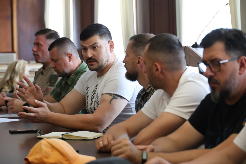 Низка людей сидять за довгим столом у залі засідань під час обговорення, де центральною фігурою є чоловік, який активно жестикулює. Чоловік у центрі кадру має коротке темне волосся, вуса та невелику борідку, одягнений у білу футболку з чорним принтом на грудях. Він емоційно говорить, широко розвівши долоні перед собою. Праворуч і ліворуч від нього вздовж столу сидять інші учасники зустрічі, переважно чоловіки, які слухають його або дивляться в бік промовця. Людина на передньому плані справа носить окуляри та має бороду. На столі перед присутніми лежать відкриті блокноти, ручки та смартфони, а на ближньому краї помітно помаранчеву кепку. Тло розмите, з лівого боку видно світлі вікна, що створюють бічне освітлення.