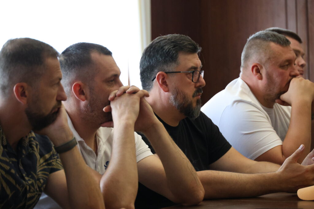 П’ятеро чоловіків сидять у ряд за столом під час «круглого столу». Вони зафіксовані у профіль та напівпрофіль. Усі присутні зосереджено дивляться праворуч, де, ймовірно, виступає промовець. Тло кімнати виконане у темних дерев'яних тонах, а зліва падає м'яке світло з вікна.