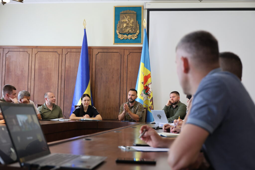 Загальний план офіційного заходу в залі засідань, де учасники сидять за великим дерев'яним столом. У центрі кадру сидить заступник голови Київської обласної державної адміністрації, ветеран Іван Добровольський, який говорить і тримає руки зчепленими перед собою. Ліворуч від нього сидять жінка-поліцейська у чорній формі та чоловік у зеленій футболці-поло, які уважно слухають промовця. Праворуч сидить чоловік у військовому одязі кольору хакі перед відкритим ноутбуком. На задньому плані між учасниками встановлено державний прапор України та прапор Київської області. Справа на передньому плані видно розмитий силует іншого учасника засідання, який робить записи в блокноті, а ліворуч на столі стоїть ноутбук, повернутий екраном до камери.