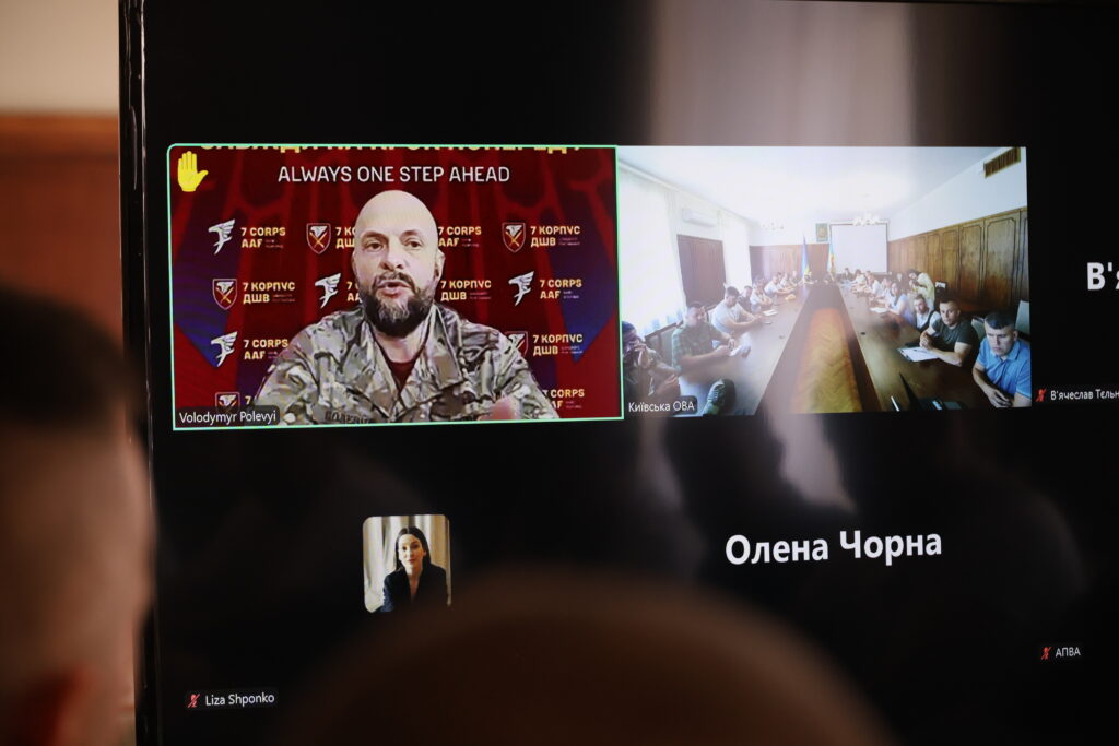 На фото зображено: великий монітор у залі засідань, на якому транслюється онлайн-конференція через Zoom або схожу програму для відеодзвінків.
